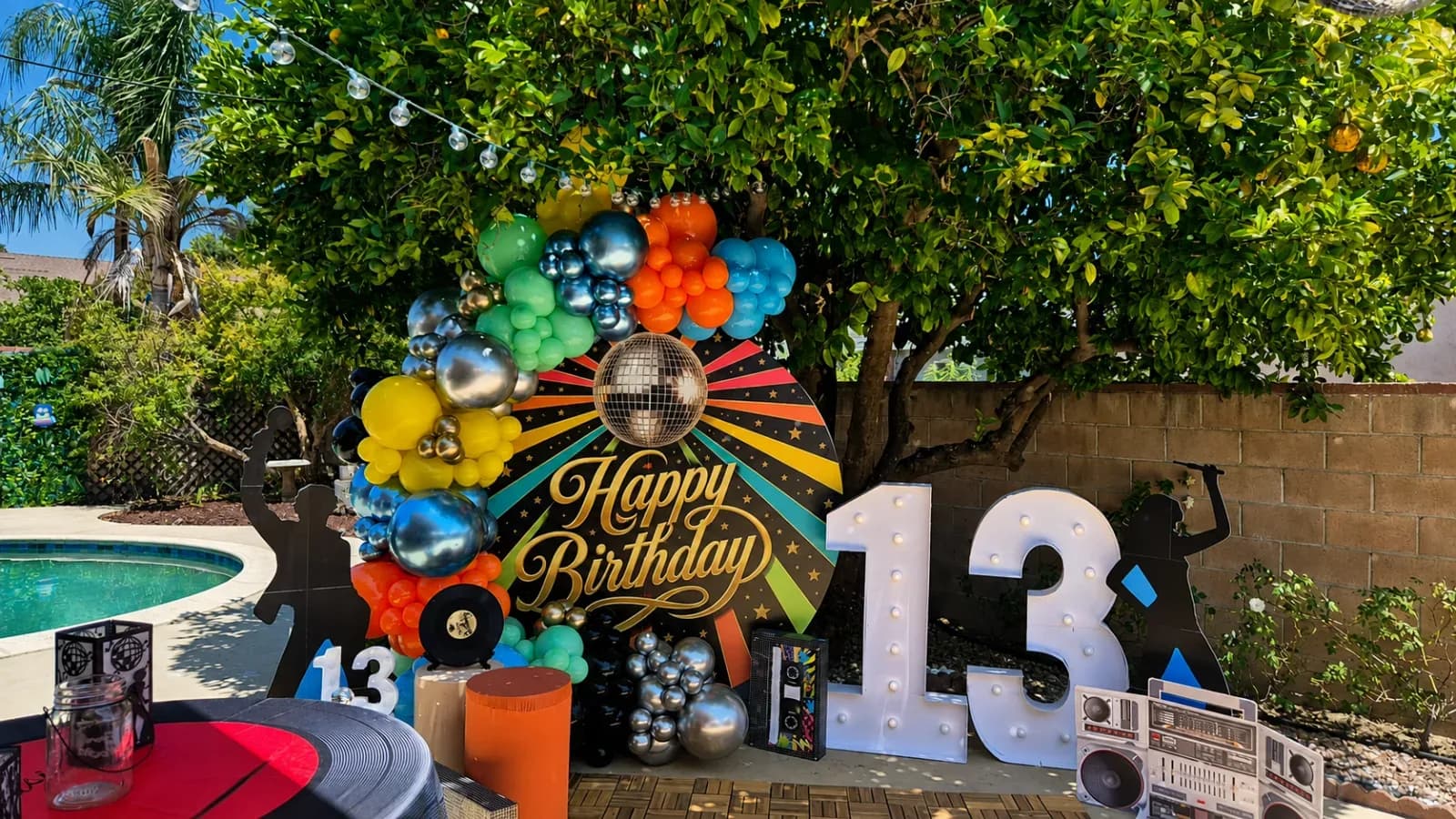 Decoración colorida con globos para cumpleaños de 13 años, fondo estilo disco retro, números iluminados, bola de discoteca y accesorios de los años 80 en Los Ángeles.