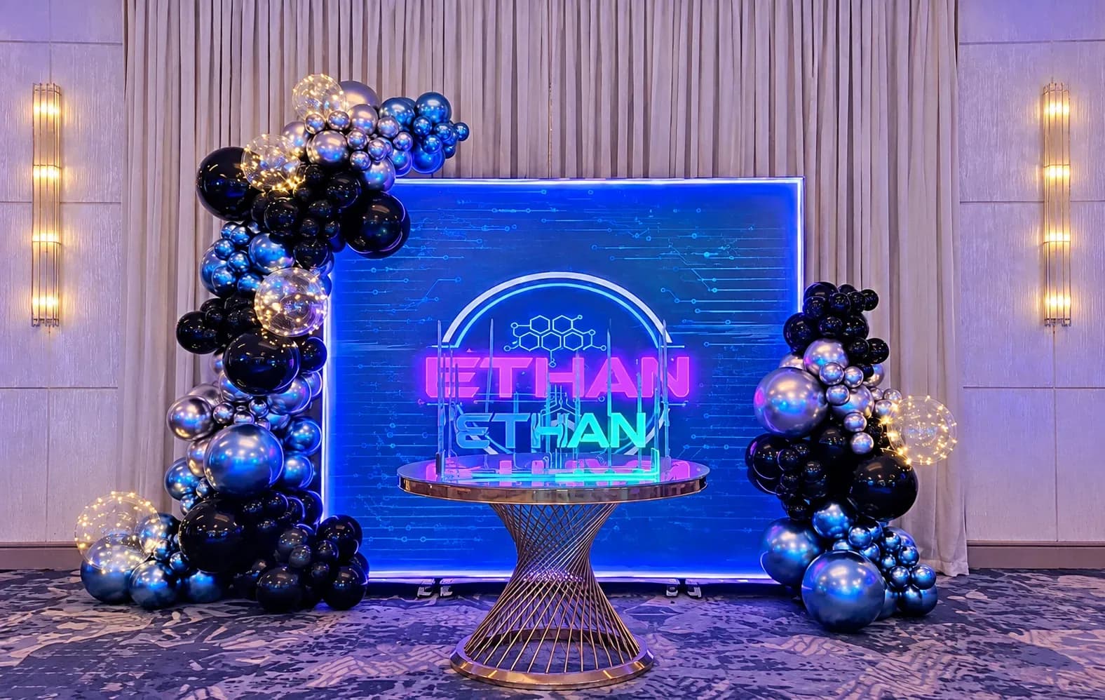 Decoración de Bar Mitzvah para niño en Los Ángeles con fondo azul personalizado e iluminado, guirnaldas orgánicas de globos negros, azul cromado y plateado metálico, globos transparentes con luces LED, diseño estilo neón y mesa dorada.